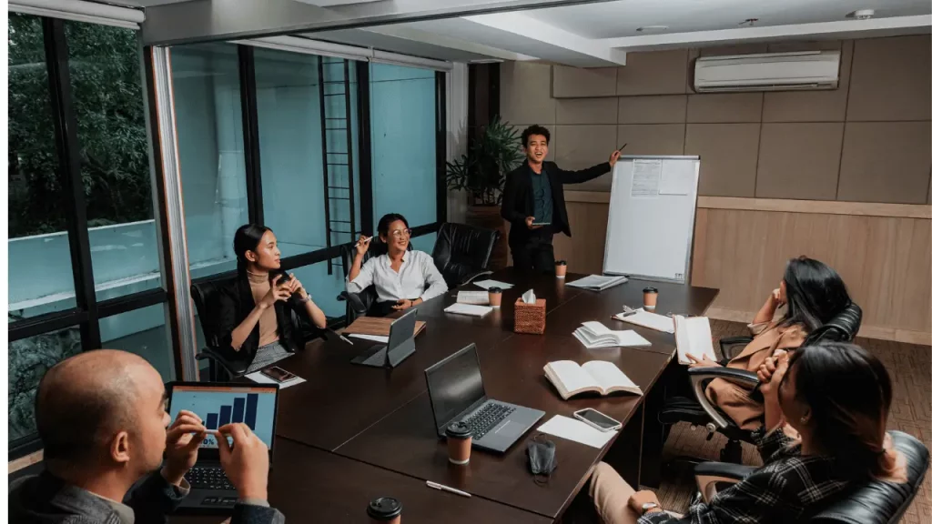 Homem em pé ao lado de um clip art, falando para uma equipe de trabalho que está sentada em uma mesa. Há homens e mulheres na equipe.