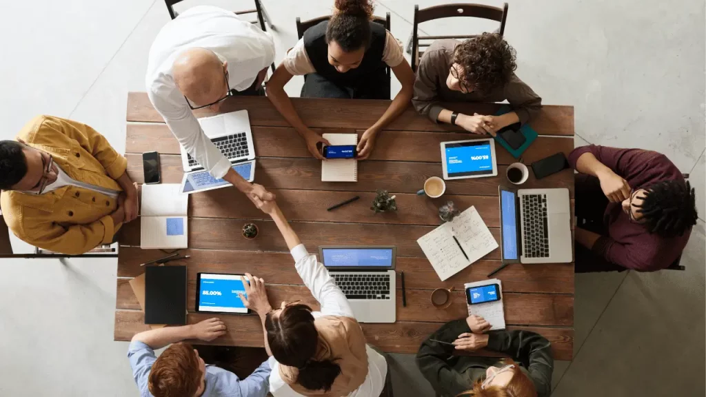 Equipe de trabalho reunida em uma grande mesa de madeira, com notebooks abertos, blocos e tablets. Há homens e mulheres na foto, sendo cinco pessoas brancas e uma mulher negra.