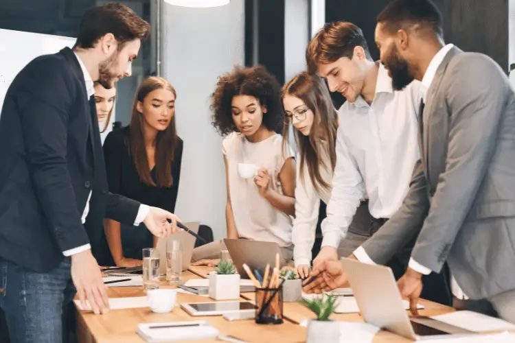 Equipe de trabalho reunida em volta de uma mesa com notebook aberto e tablets. As pessoas analisam relatórios que estão na mesa.