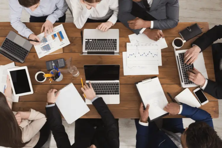 Imagem mostra uma equipe reunida em uma mesa grande de madeira, com muitos papéis com dados e notebooks abertos.