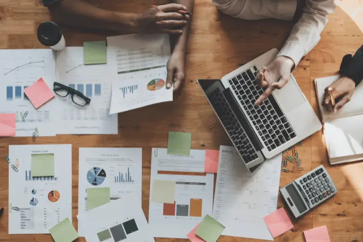 Pessoas reunidas em uma mesa, que em cima há muitos dados em papel e um notebook aberto.