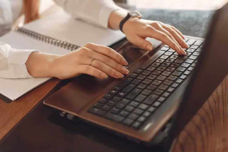 Imagem de mãos femininas digitando em um notebook e um caderno em frente ao computador
