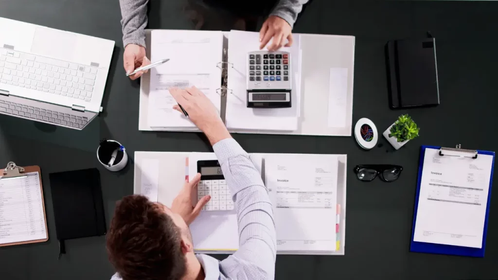 Dois homens em uma mesa, um de frente para o outro, com documentos e calculadoras entre eles. Os dois são brancos e usam roupas sociais.