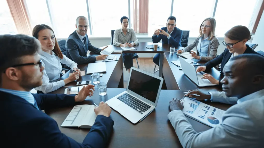 Reunião em uma empresa, com muitas pessoas sentadas em volta de uma grande mesa. Em cima da mesa há documentos e um notebook aberto. A maioria das pessoas são homens e brancos, exceto um que é negro. Também há três mulheres brancas. Todas as pessoas vestem roupas sociais. 