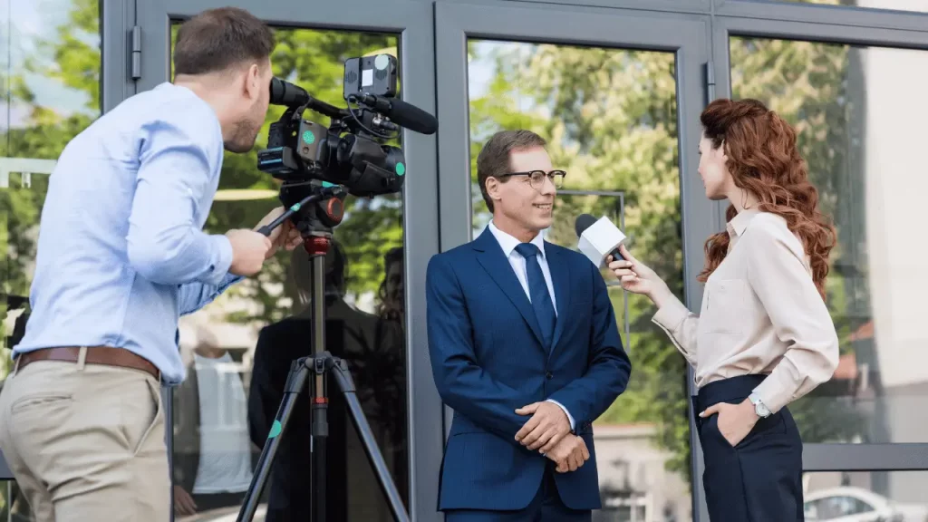 Na imagem, uma repórter de TV entrevistando um homem. Ela é branca e veste camisa branca e calça no estilo social. O entrevistado é um homem branco e veste terno.
