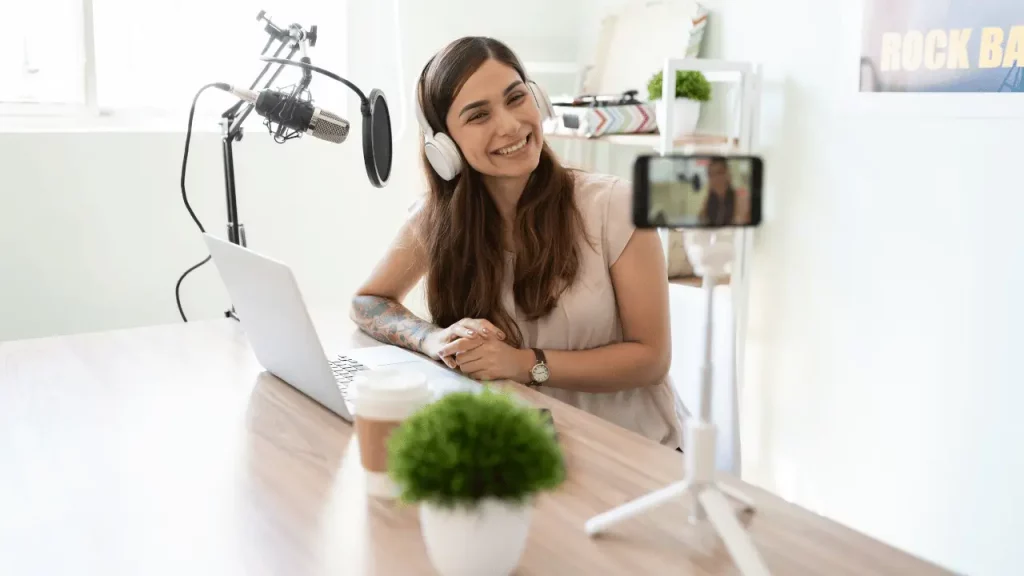 Mulher jovem e branca, olhando para um celular que está filmando ela, com um notebook em sua frente e microfone também.
