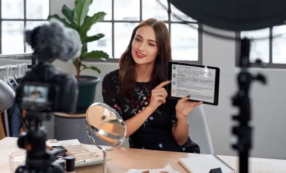 Mulher jovem, branca, falando para uma câmera e mostrando algo em um tablet.