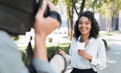 Imagem mostra uma repórter de TV, com o microfone na mão e um câmera filmando ela. A mulher é negra e veste camisa social branca.