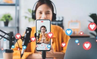 imagem de uma influenciadora digital. Em primeiro plano está um celular com a imagem dela e ela atrás, em segundo plano. Emojis de corações também aparecem na tela