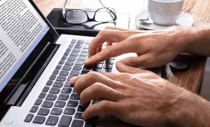 Homem digitando em um notebook, que na tela mostra um bloco de texto. Aparece apenas as mãos do homem. Ele é branco.