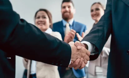 Dois homens de terno apertando as mãos em primeiro plano. Ao fundo, um homem e duas mulheres vestindo roupas socias. Todas as pessoas da foro são brancas.