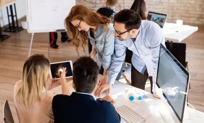 Pessoas reunidas em uma mesa de trabalho, analisando algo na tela de um tablet. Há duas mulheres e dois homens e todos são brancos e vestem roupas sociais.