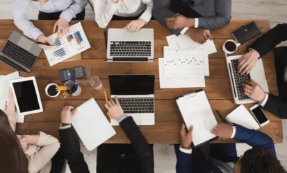 Imagem mostra uma equipe reunida em uma mesa grande de madeira, com muitos papéis com dados e notebooks abertos.