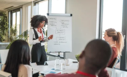 Mulher em pé, ao lado de um clip art, falando pra um grupo de trabalho, composto por três pessoas, sendo duas mulheres brancas e um homem negro. Ela é negra.
