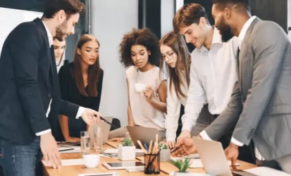 Equipe de trabalho reunida em volta de uma mesa com notebook aberto e tablets. As pessoas analisam relatórios que estão na mesa.